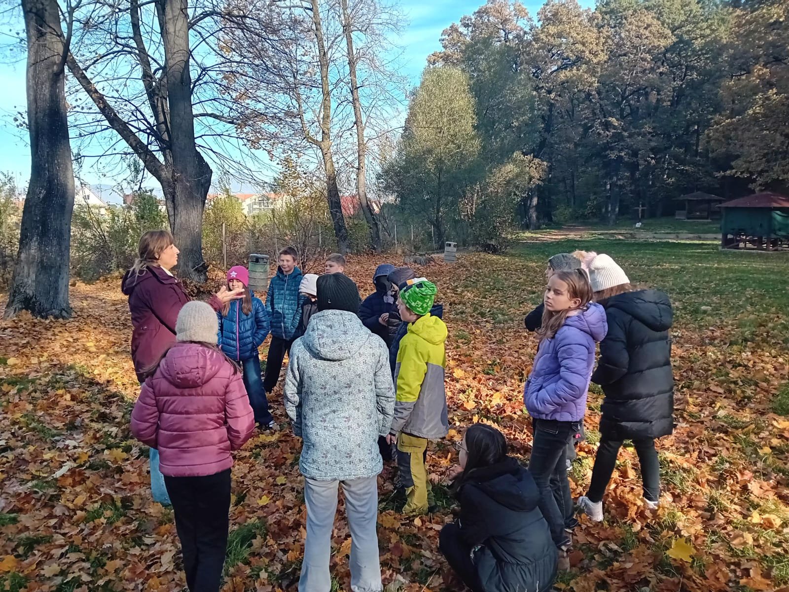 Grup de elevi îmbrăcați de toamnă, ascultând și discutând despre recunoștință alături de un formator ACPEIO, în mijlocul frunzelor căzute.