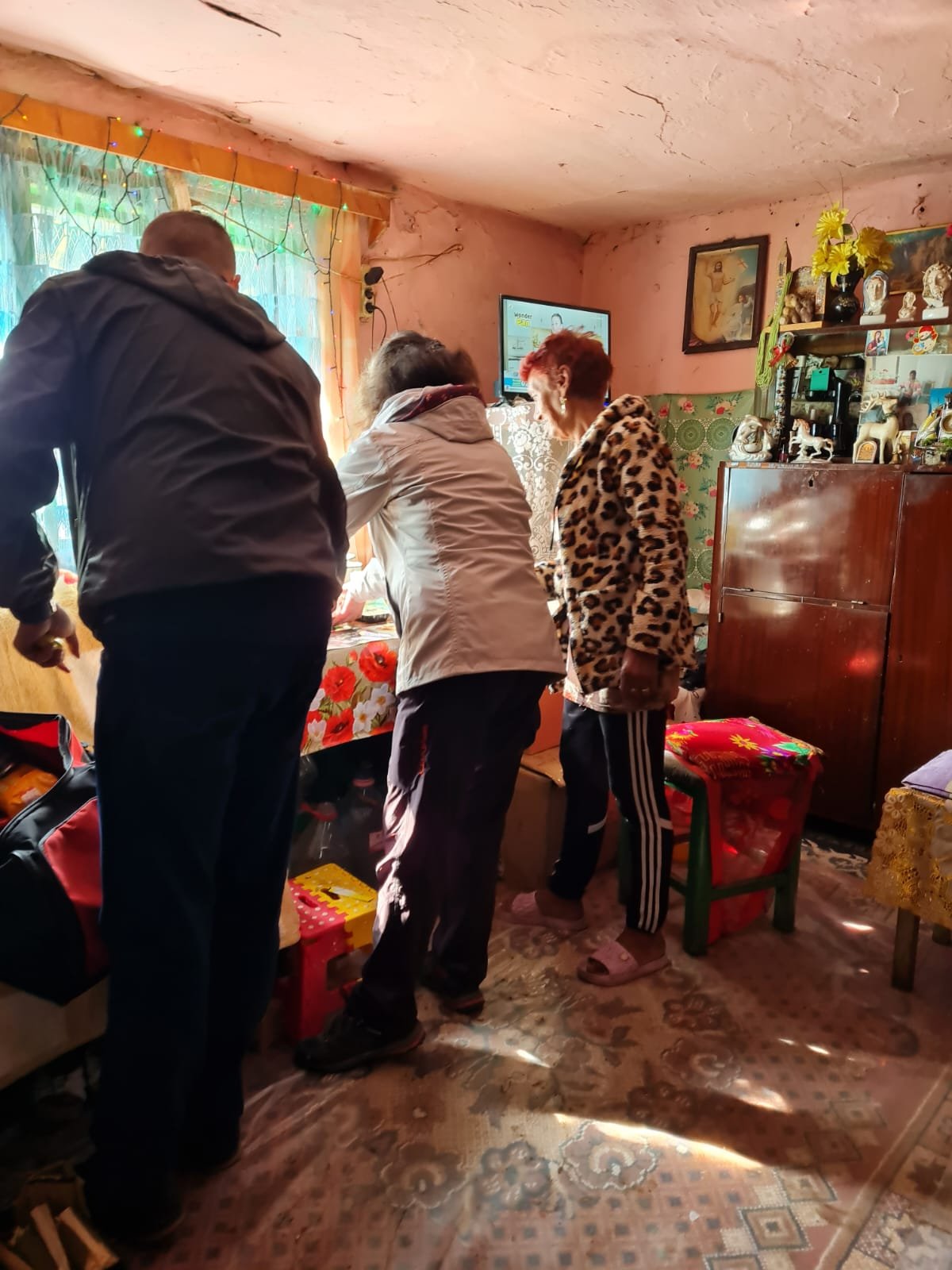 Trei voluntari ai proiectului „O Mână Întinsă în Cătunele din Apuseni” aranjează pachete cu alimente pe o masă într-o locuință modestă a unei persoane vârstnice.
