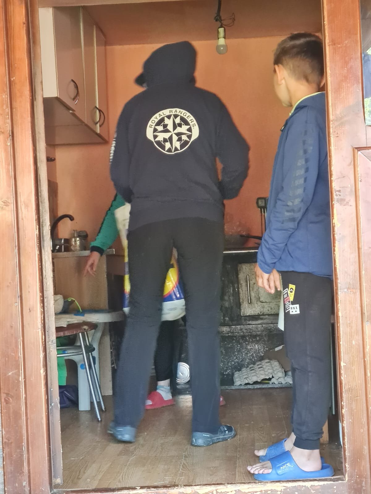 Voluntar îmbrăcat cu hanorac Royal Rangers și un copil stau în pragul unei case din Apuseni, în timp ce în interior sunt descărcate sacoșe cu alimente și provizii.
