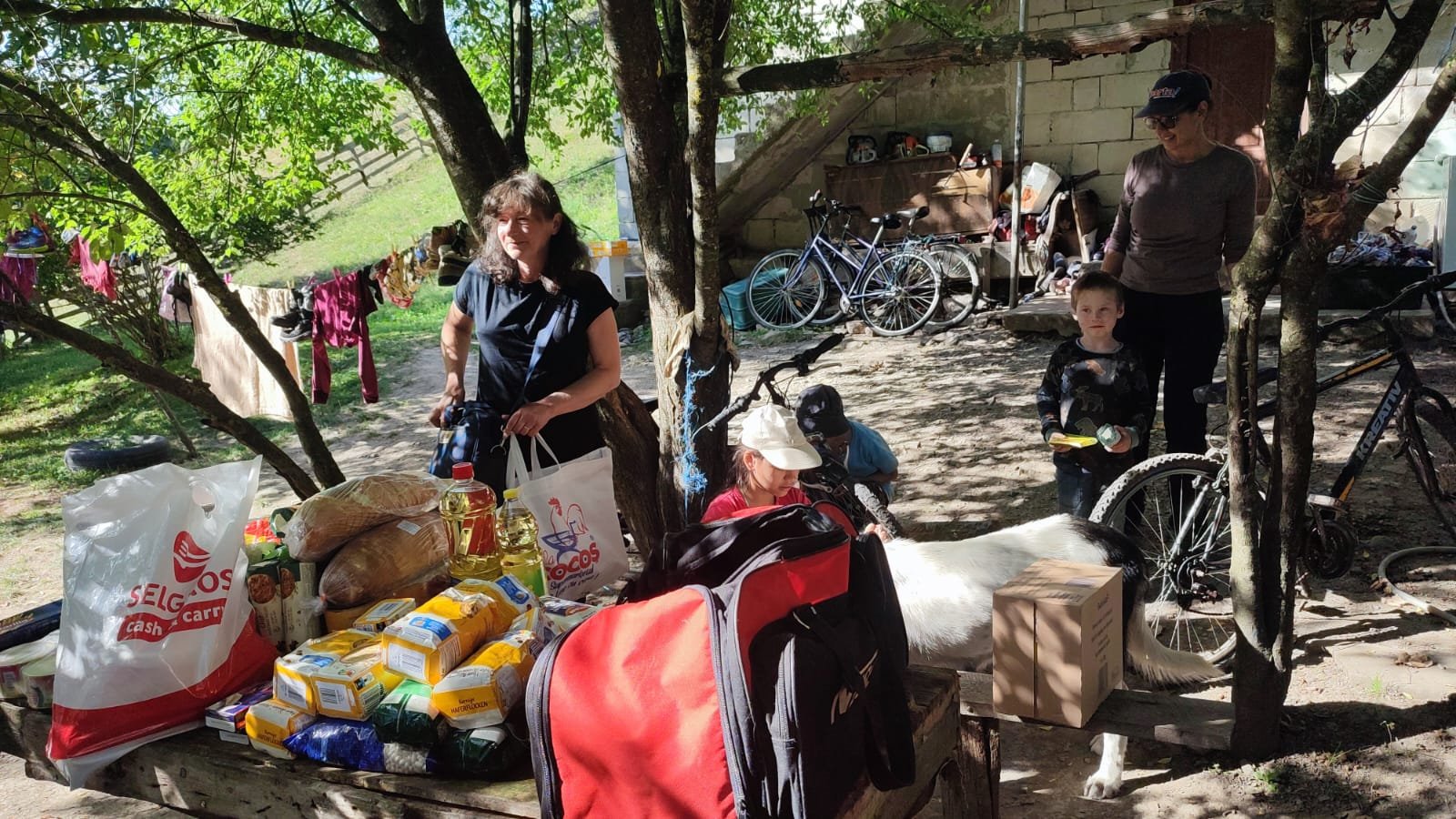 Voluntari și copii dintr-o familie din Apuseni adunați în curte, lângă o masă plină cu pâine, ulei, făină și alte alimente oferite prin proiectul „O Mână Întinsă în Cătunele din Apuseni”, în parteneriat cu Asociația CPEIO.