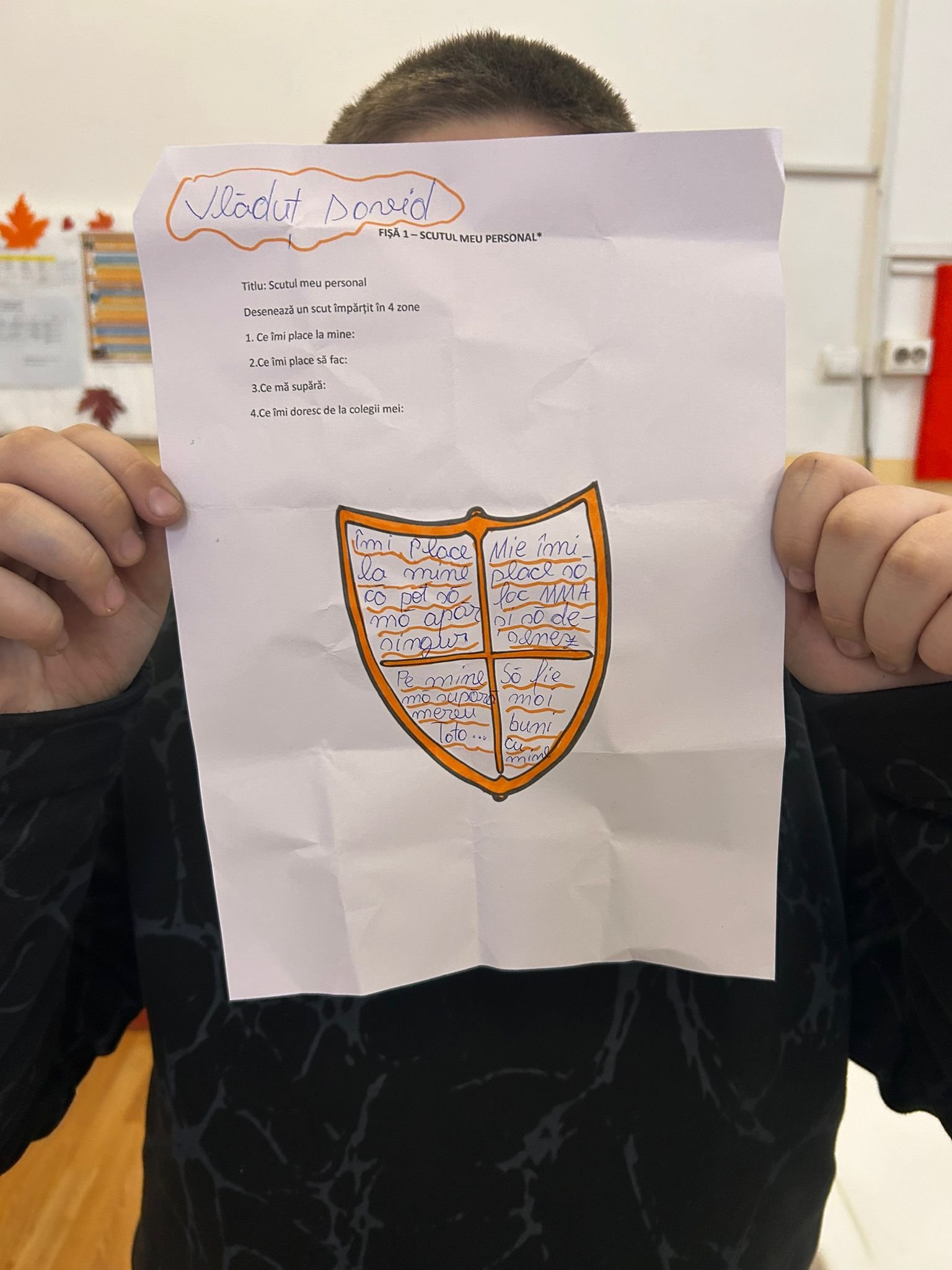 Elev ține un scut colorat pe care a scris ce îi place la el, ce îl întristează și cum și-ar dori să fie relația cu colegii săi.