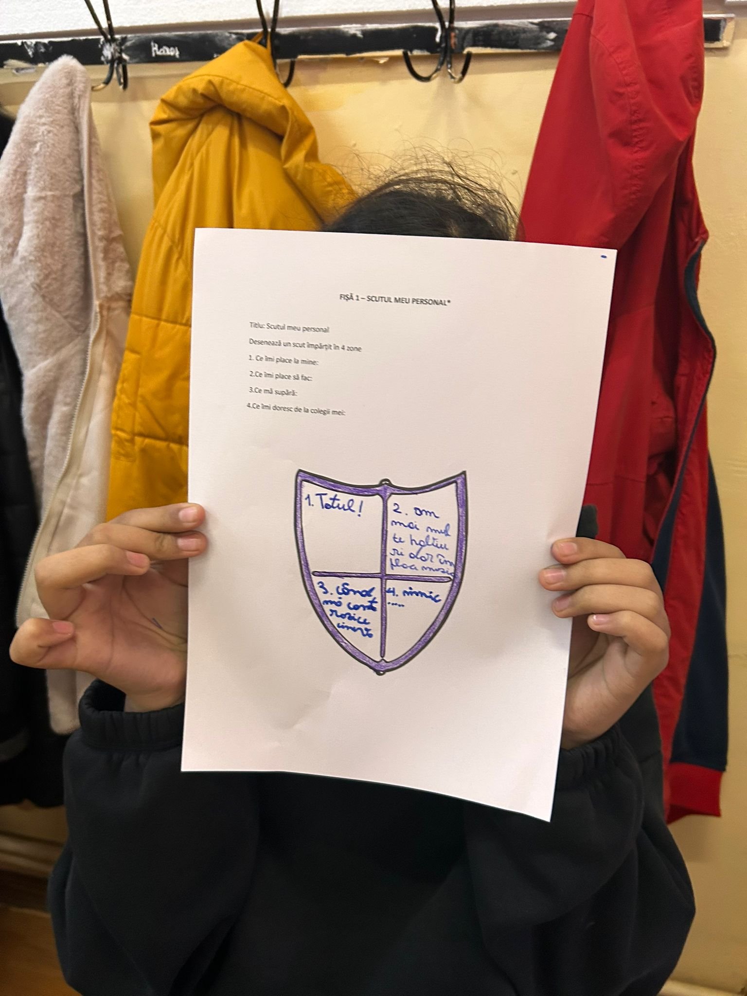 Elev care își acoperă fața cu o fișă „Scutul meu personal”, pe care sunt scrise ce îi place, ce îl supără și ce își dorește de la colegi, în timpul atelierului ACPEIO.