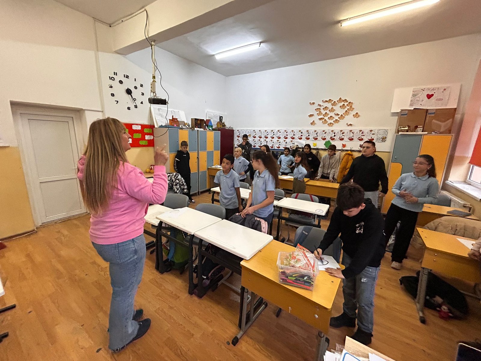 Facilitator ACPEIO în hanorac roz coordonează un exercițiu interactiv cu elevii clasei a V-a, care stau în picioare printre bănci și lucrează la fișele lor în cadrul programului „Scânteia schimbării”.