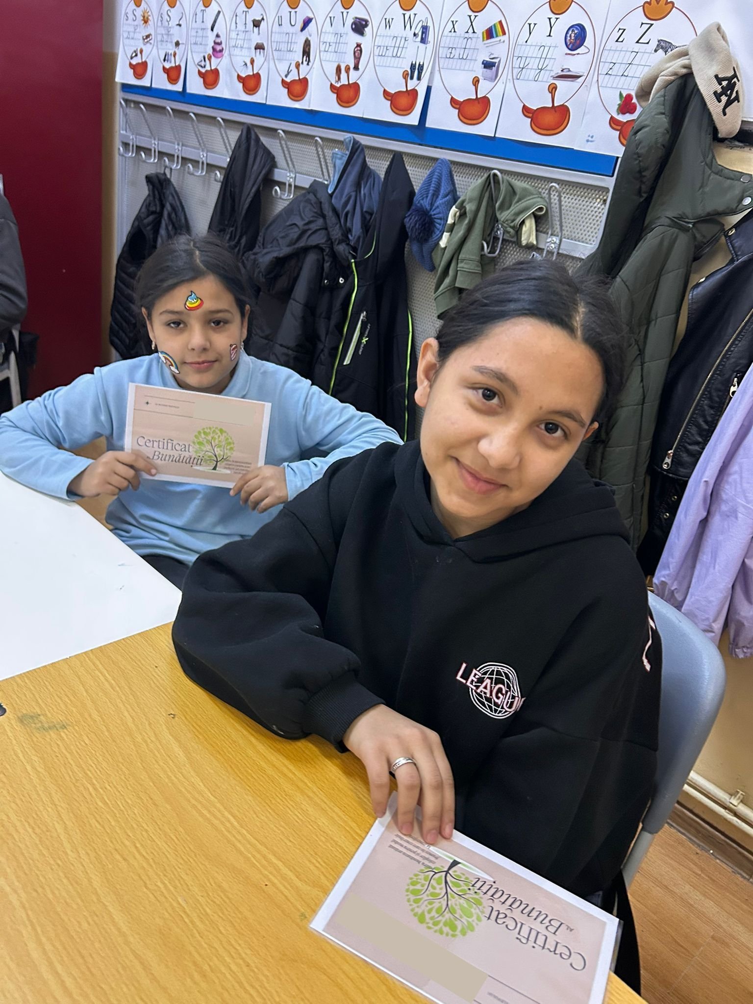 Două eleve stau în bancă și țin în mâini „Certificatul Bunătății”, la finalul unei sesiuni de dezvoltare socio-emoțională organizate de ACPEIO.