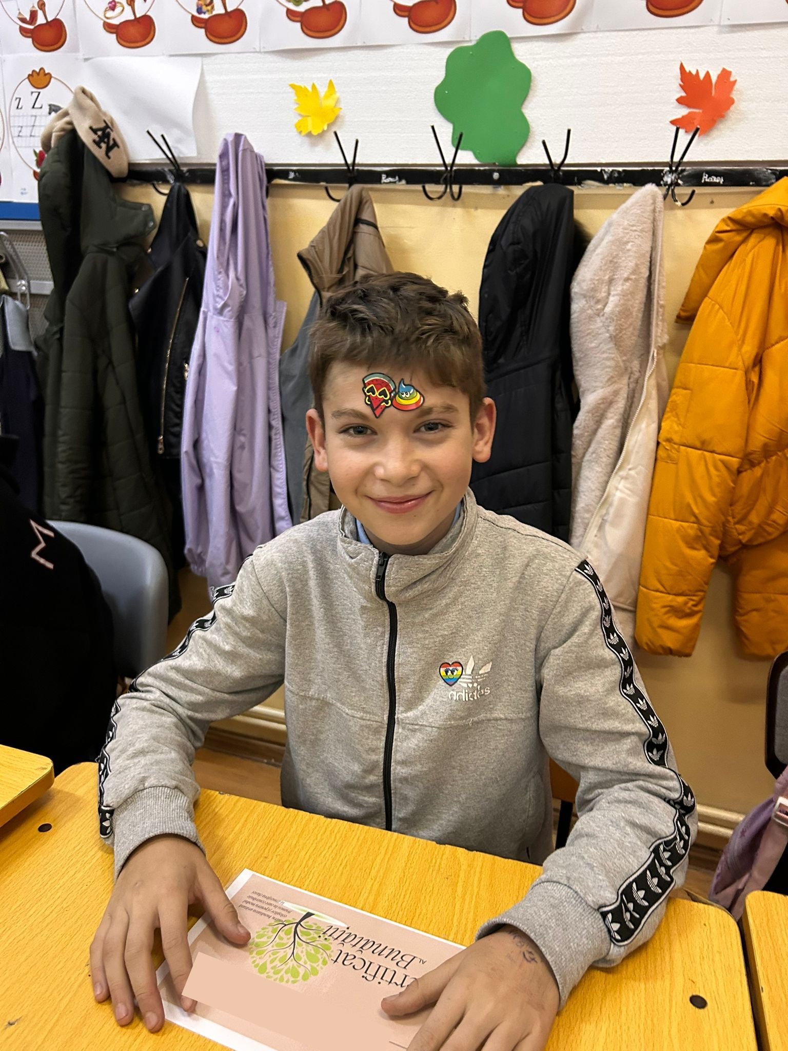 Elev zâmbitor la bancă, cu stickere colorate pe frunte și cu „Certificatul Bunătății” în față, ca parte a activităților ACPEIO pentru dezvoltare emoțională.