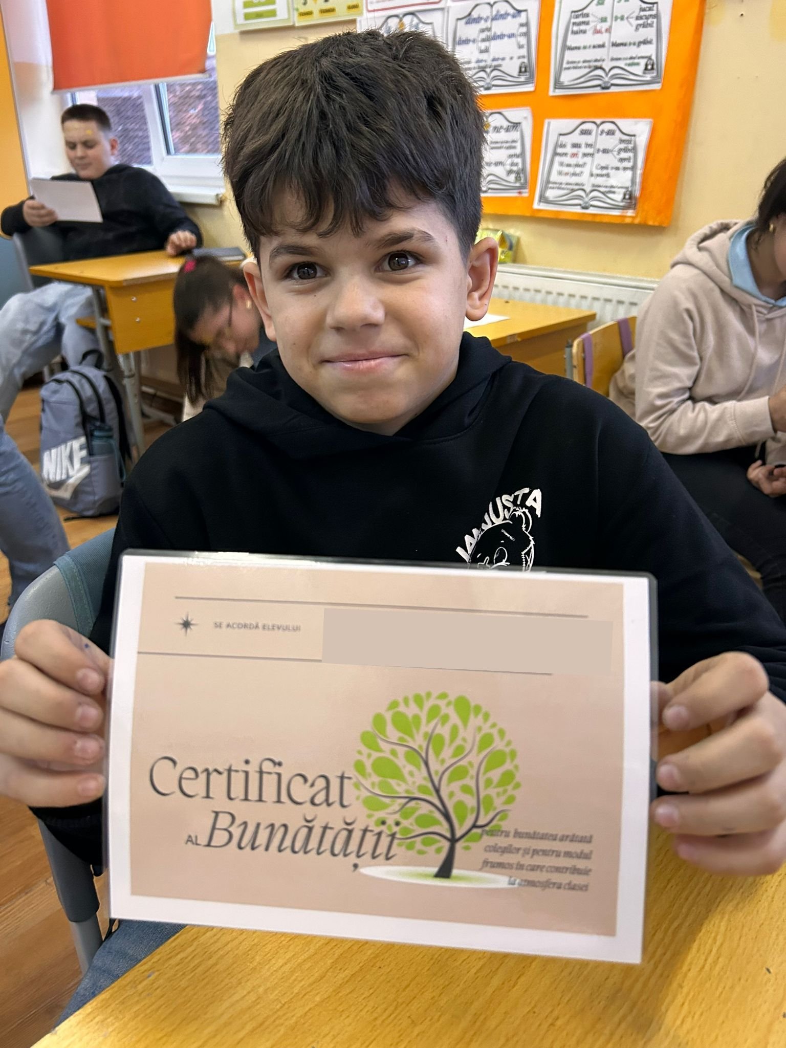 Elev ține în mâini „Certificatul Bunătății” și zâmbește către aparat, în sala de clasă, la o activitate ACPEIO dedicată cultivării bunătății și respectului.