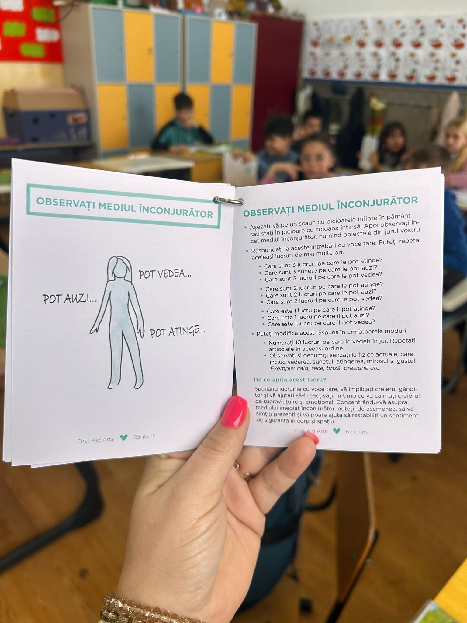 Caiet First Aid Arts deschis la exercițiul „Observați mediul înconjurător”, ținut în mână, cu elevi care lucrează în fundal în sala de clasă.