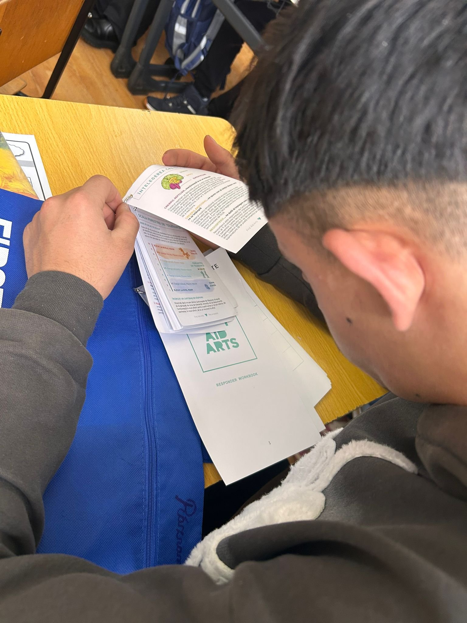 Elev răsfoind caietul „Responder Workbook” First Aid Arts, așezat la bancă, cu materialele de lucru pe masă.