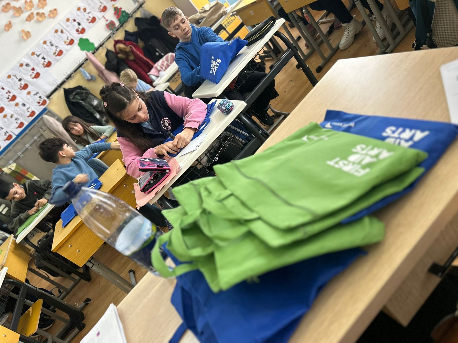 Vedere din clasă cu elevi care lucrează și sacoșe verzi și albastre First Aid Arts așezate pe catedră în prim-plan.