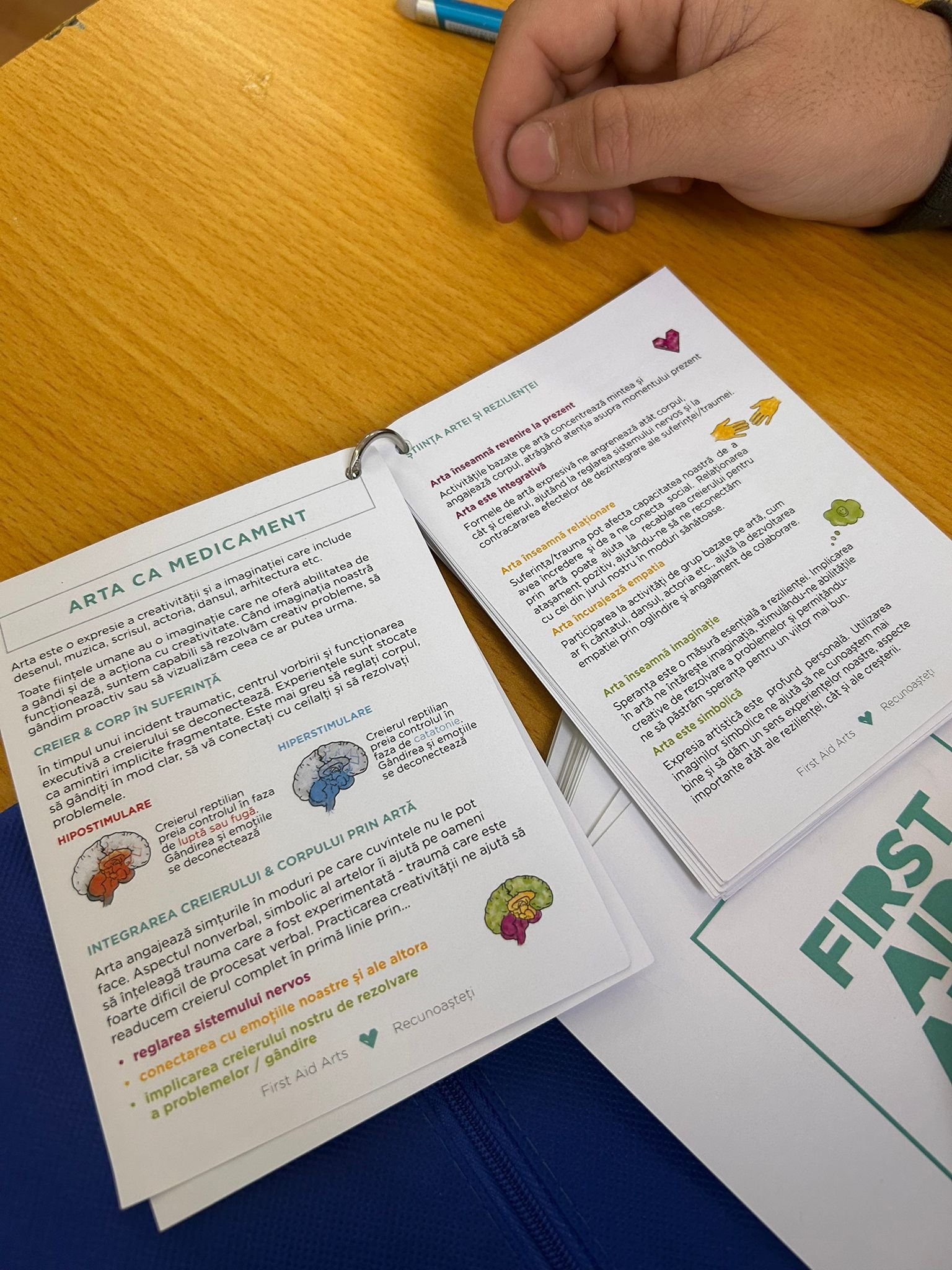 Prim-plan cu fișele educaționale First Aid Arts despre rolul artei în reglarea emoțiilor și creșterea rezilienței, pe banca unui elev.