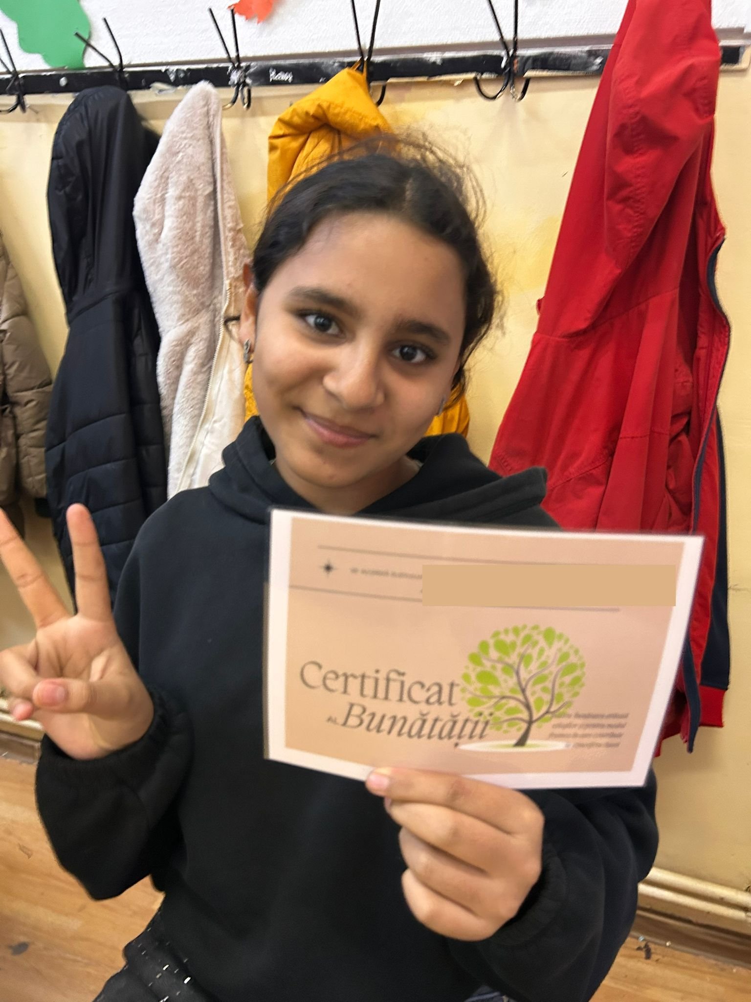 Elevă zâmbitoare, făcând semnul păcii și ținând în mână „Certificatul Bunătății”, la activitățile ACPEIO din programul „Scânteia schimbării”.