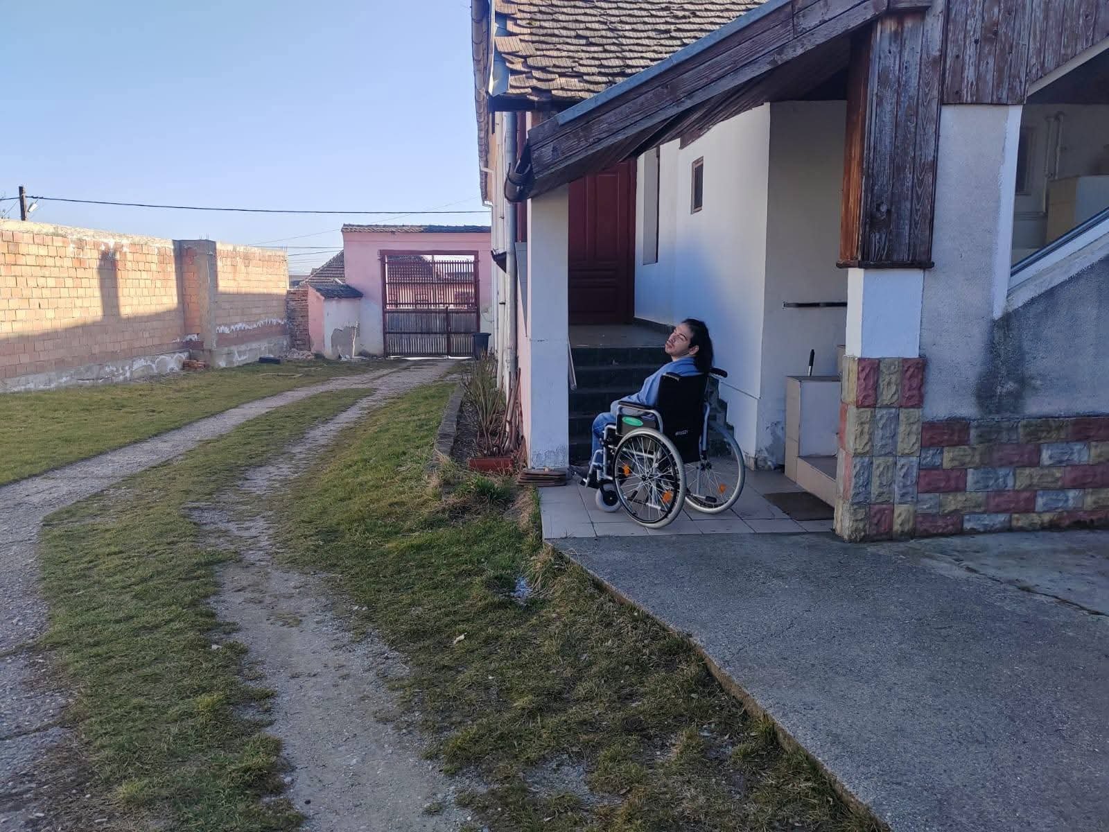 Persoană în scaun rulant în fața unei case din Rotbav, spațiu propus pentru tabără de creație și dezvoltare personală pentru persoane cu dizabilități
