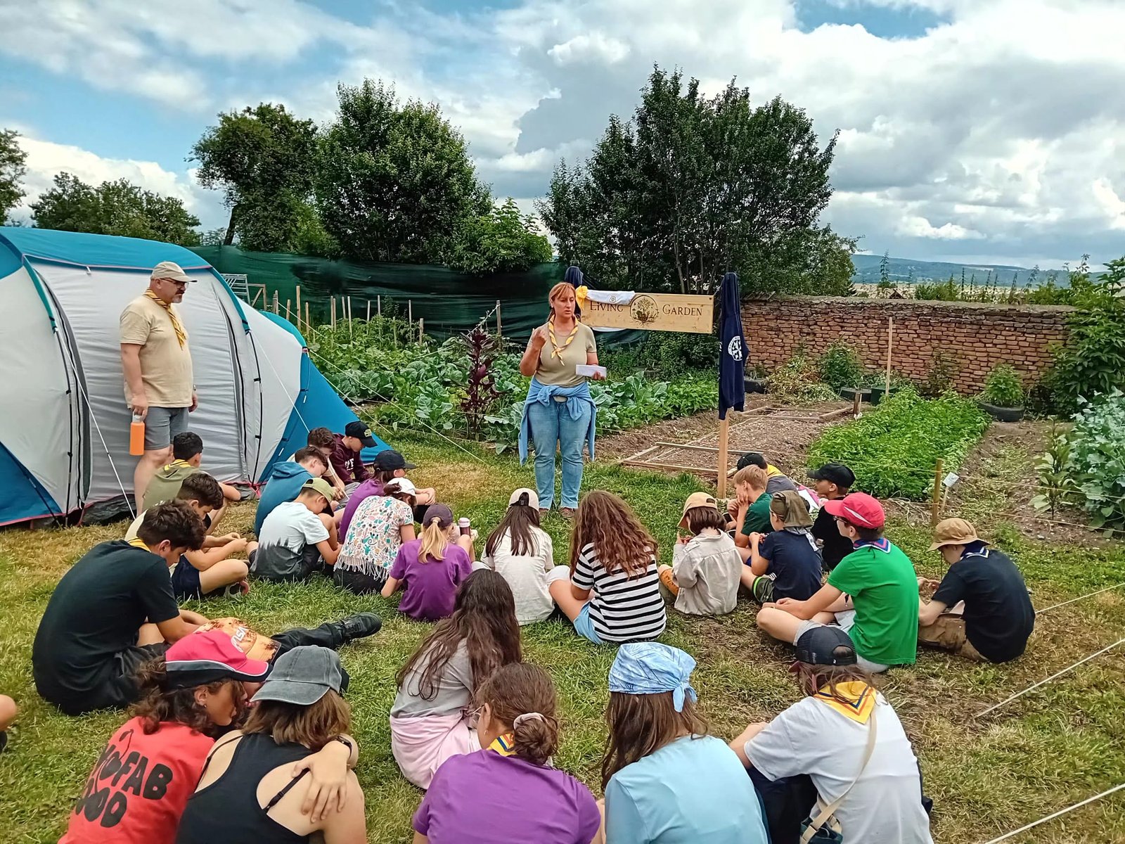 Grup de copii așezați pe iarbă ascultând o coordonatoare la un atelier educațional în aer liber, lângă straturile de legume din Grădina Vie – Living Garden
